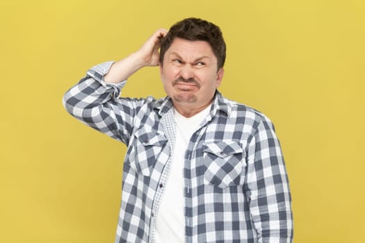 Portrait of thoughtful attractive middle aged man in casual checkered shirt, keeps hand on his head, thinking, trying to remember something. Indoor studio shot isolated on yellow background.