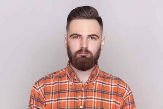 Portrait of good-looking serious bearded man standing looking at camera with strict bossy facial expression, wearing checkered shirt. Indoor studio shot isolated on gray background.