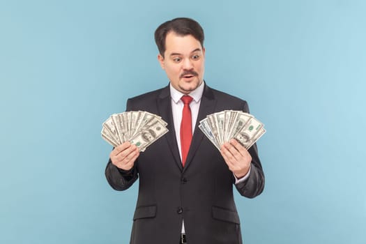 Portrait of rich man with mustache standing counting money., holding big fans of dollar banknotes, wearing black suit with red tie. Indoor studio shot isolated on light blue background.
