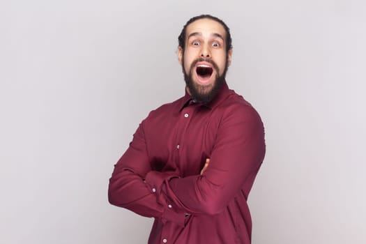Portrait of amazed surprised man with dark hair and beard in red shirt standing with crossed arms, looking at camera with big eyes. Indoor studio shot isolated on gray background.