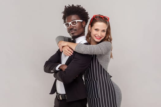 Portrait of attractive happy woman and man in white glasses standing together, wife hugging her self-confident husband. Indoor studio shot isolated on gray background.