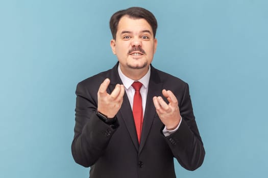 Portrait of angry aggressive man with mustache standing with raised arms, expressing anger and hate, wearing black suit with red tie. Indoor studio shot isolated on light blue background.