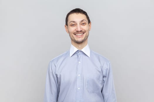 Portrait of handsome smiling joyful man standing looking at camera with toothy smile, expressing happiness, wearing light blue shirt. Indoor studio shot isolated on gray background.