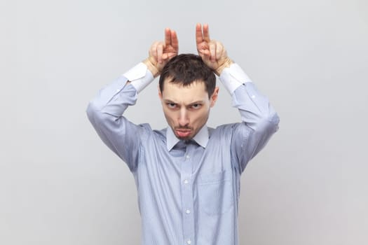 Portrait of angry serious aggressive man standing and showing bully horns, attacks, looks crazy and dangerous, wearing light blue shirt. Indoor studio shot isolated on gray background.