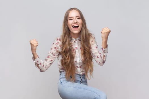 I am champion. Portrait of woman with wavy blond hair screaming for joy and raising high her fists, celebrating successful winning, incredible victory. Indoor studio shot isolated on gray background.