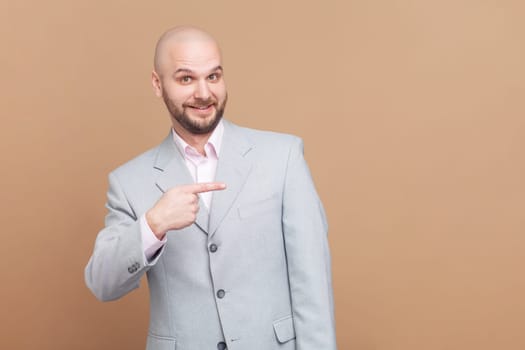 Portrait of smiling satisfied bald bearded man standing pointing at advertisement area, copy space for promotion, wearing gray jacket. Indoor studio shot isolated on brown background.