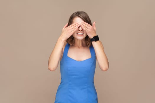 Don't want to look at this. Portrait of scared woman with wavy hair standing, covering eyes with hand, refusing to watch, wearing blue dress. Indoor studio shot isolated on light brown background.