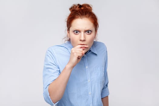 Portrait of redhead woman wearing blue shirt biting finger with panicked terrified face, having depression and nervous problem at work. Indoor studio shot isolated on gray background.