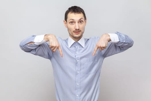 Portrait of serious strict young adult man standing pointing both index fingers down, saying here and right now, wearing light blue shirt. Indoor studio shot isolated on gray background.