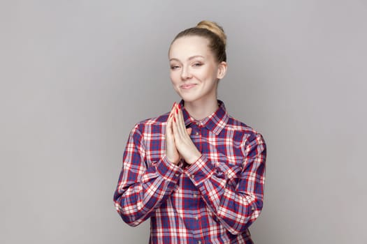 Portrait of sly attractive woman with bun hairstyle keeps palms together, planning devil plan or prank, wearing checkered shirt. Indoor studio shot isolated on gray background.