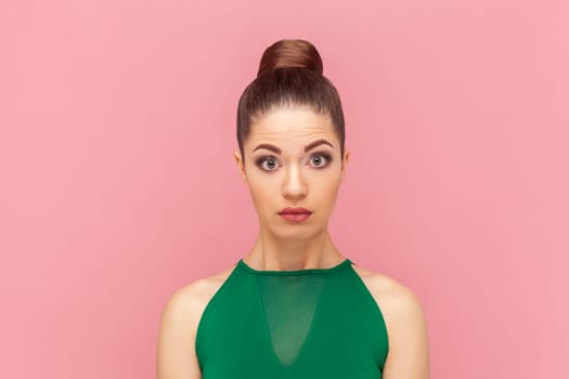 Portrait of attractive shocked amazed woman standing and looking at camera with big eyes, sees something astonishing, wearing green dress. Indoor studio shot isolated on pink background.