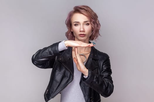 Portrait of adorable sad woman with short hairstyle standing showing time out gesture, needs more time for her work, wearing black leather jacket. Indoor studio shot isolated on grey background.