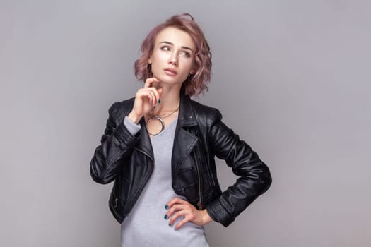 Portrait of pensive thoughtful woman with short hairstyle standing holding her chin, thinking about her work, wearing black leather jacket. Indoor studio shot isolated on grey background.