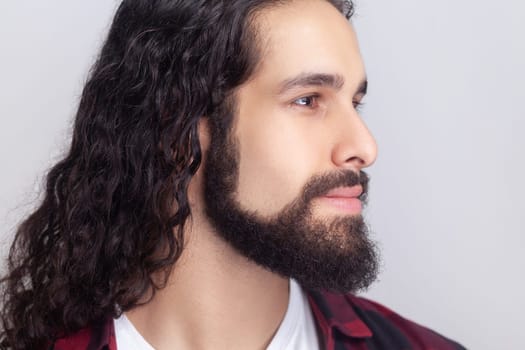Closeup portrait of calm handsome bearded man with long curly hair in checkered red shirt looking away with smile, expressing positive emotions. Indoor studio shot isolated on gray background.