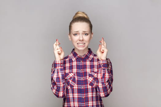 Portrait of hopeful attractive woman with bun hairstyle standing with clenched teeth, crossing fingers, praying for good luck, wearing checkered shirt. Indoor studio shot isolated on gray background.