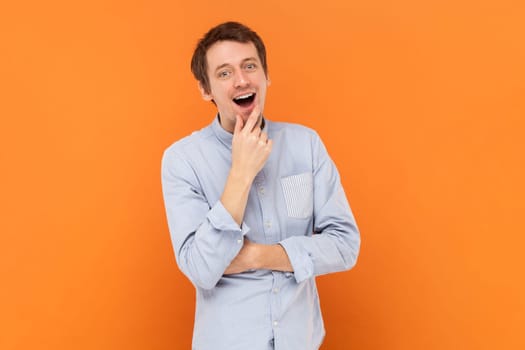 Portrait of excited amazed young adult man standing with hand on chin, having good idea, looking at camera, wearing light blue shirt. Indoor studio shot isolated on orange background.