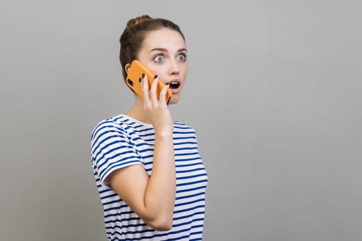 Side view of astonished woman wearing striped T-shirt talking via cell phone, hearing shocking news, looking way with open mouth. Indoor studio shot isolated on gray background.