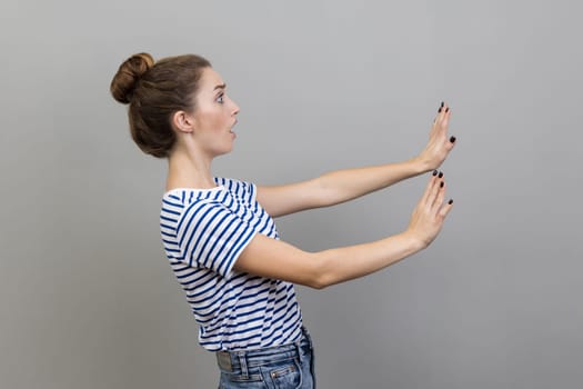 Side view portrait of scared young adult woman wearing striped T-shirt gesturing stop with palms and looking surprised with frightened eyes. Indoor studio shot isolated on gray background.