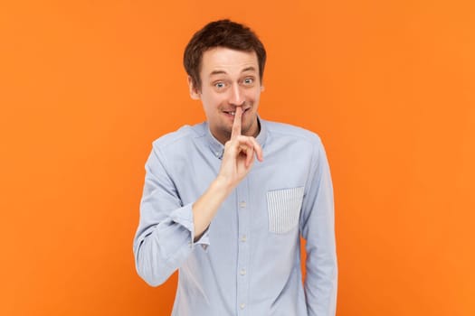 I won't tell ever. Portrait of funny positive optimistic man looking at camera and showing shh gesture, wearing light blue shirt. Indoor studio shot isolated on orange background.