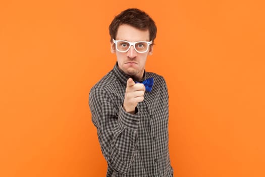 Self assured serious man nerd points index finger at camera chooses you looks directly at camera, wearing shirt with blue bow tie and white glasses. Indoor studio shot isolated on orange background.