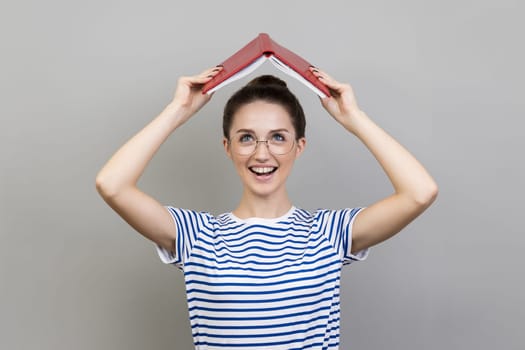 Portrait of woman wearing striped T-shirt and glasses holding opened book on head, looking at camera with smile, expressing positive emotions. Indoor studio shot isolated on gray background.