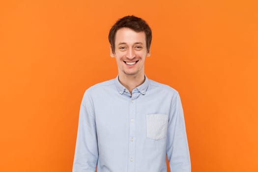 Portrait of overjoyed positive happy man standing looking at camera with toothy smile, expressing happiness, wearing light blue shirt. Indoor studio shot isolated on orange background.