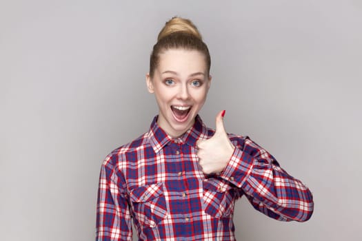 Portrait of amazed excited woman with bun hairstyle keeps thumb raised, being in good mood, shows her agreement, wearing checkered shirt. Indoor studio shot isolated on gray background.