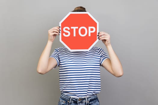 Portrait of woman wearing striped T-shirt hiding her face behind stop sign, afraid talking about domestic violence, scared victim. Indoor studio shot isolated on gray background.