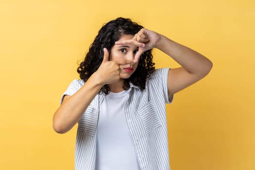 Portrait of woman with dark wavy hair making frame gesture with her fingers as she looks through camera, composition of photograph. Indoor studio shot isolated on yellow background.
