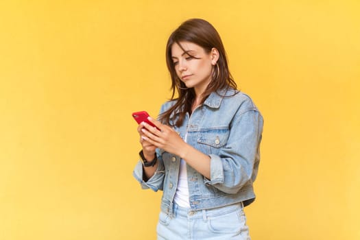 Side view portrait of serious concentrated woman wearing denim jacket using cell phone, chatting with somebody with attentive expression. Indoor studio shot isolated on yellow background.