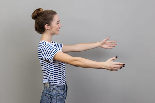 Come into my arms. Side view portrait of woman wearing striped T-shirt stretching hands and smiling broadly, going to embrace, share love. Indoor studio shot isolated on gray background.