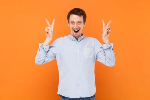 Portrait of satisfied optimistic young adult man standing showing v sign, looking at camera, celebrating victory, wearing light blue shirt. Indoor studio shot isolated on orange background.
