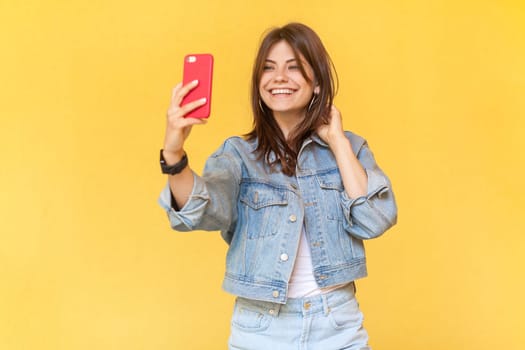 Portrait of charming pretty smiling woman blogger wearing denim jacket holding mobile phone in hands, making selfie or broadcasting livestream. Indoor studio shot isolated on yellow background.