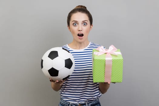 Portrait of surprised woman wearing striped T-shirt standing looking at camera with big eyes, holding out soccer ball and green present box. Indoor studio shot isolated on gray background.