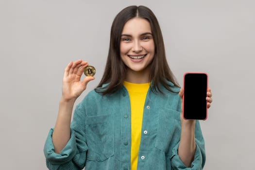 Smiling happy woman holding gold bitcoin and smartphone with empty screen for advertisement or promotion, wearing casual style jacket. Indoor studio shot isolated on gray background.