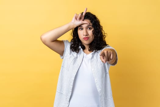 Portrait of grumpy disrespectful woman with dark wavy hair showing loser gesture and pointing finger at camera, blaming and mocking. Indoor studio shot isolated on yellow background.