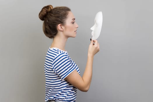 Side view portrait of beautiful dark haired woman with bun hairstyle wearing striped T-shirt standing holding and looking at white mask. Indoor studio shot isolated on gray background.