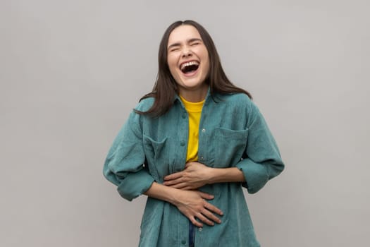 Happy woman holding her stomach and laughing out loud, chuckling giggling at amusing anecdote, sincere emotion, wearing casual style jacket. Indoor studio shot isolated on gray background.