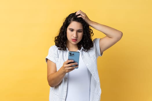 Portrait of pensive beautiful young adult woman with dark wavy hair using cell phone thinking what to answer, keeps hand on head. Indoor studio shot isolated on yellow background.