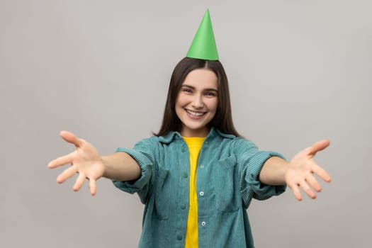 Woman with party cone keeping hands wide open to embrace, giving warm welcome, greeting and sharing love, wearing casual style jacket. Indoor studio shot isolated on gray background.