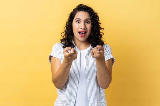 Portrait of amazed excited beautiful woman with dark wavy hair pointing directly at you, choosing something, has shocked expression. Indoor studio shot isolated on yellow background.
