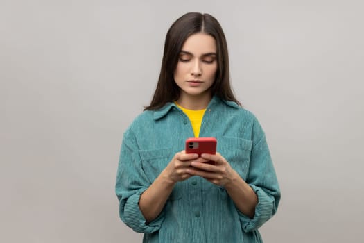 Serious pretty woman reading message on smartphone, using mobile device for communication, browsing web, wearing casual style jacket. Indoor studio shot isolated on gray background.