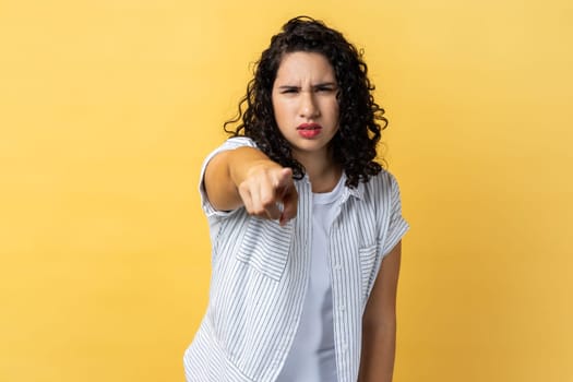 Portrait of strict woman with dark wavy hair noticing and pointing finger to camera, accusing with serious bossy face, making choice. Indoor studio shot isolated on yellow background.
