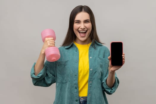 Portrait of happy excited holding pink dumbbell and blank screen smartphone in hands, satisfied with sports app, wearing casual style jacket. Indoor studio shot isolated on gray background.