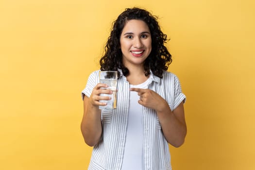Portrait of smiling happy positive beautiful woman with dark wavy hair standing with glass of water, pointing at beverage with finger. Indoor studio shot isolated on yellow background.