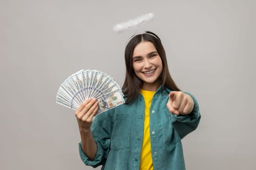 Happy angelic young woman with halo above head holding dollar banknotes and pointing to camera, choosing lottery winner, encouraging to earn big money. Indoor studio shot isolated on gray background.