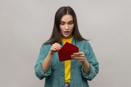 Portrait of amazed surprised woman holding red romantic envelope and looking at letter with open mouth, wearing casual style jacket. Indoor studio shot isolated on gray background.