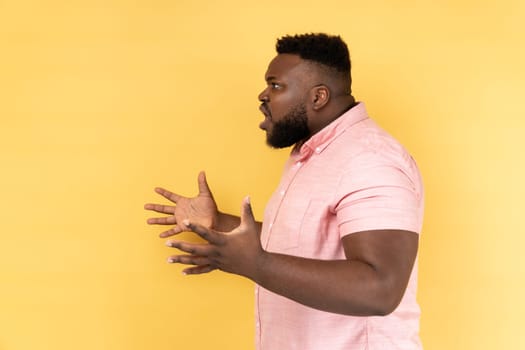 Side view of annoyed stressed man wearing pink shirt asking what do you want and spreading hands, family quarrel, misunderstanding. Indoor studio shot isolated on yellow background.