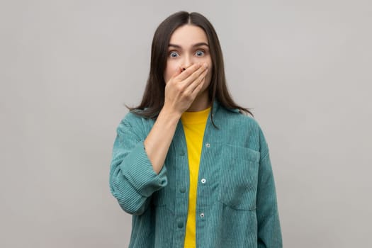 Portrait of scared shocked dark haired woman closing mouth with hand, afraid to say too much, embarrassment, wearing casual style jacket. Indoor studio shot isolated on gray background.