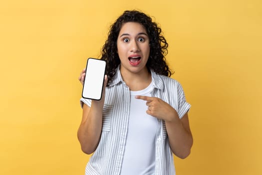 Portrait of amazed surprised woman with dark wavy hair holding pointing ar mobile phone with blank screen, mockup for advertisement. Indoor studio shot isolated on yellow background.